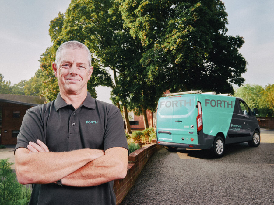 A smiling engineer wearing a FORTH® embossed polo shirt stands proudly in front of his van.